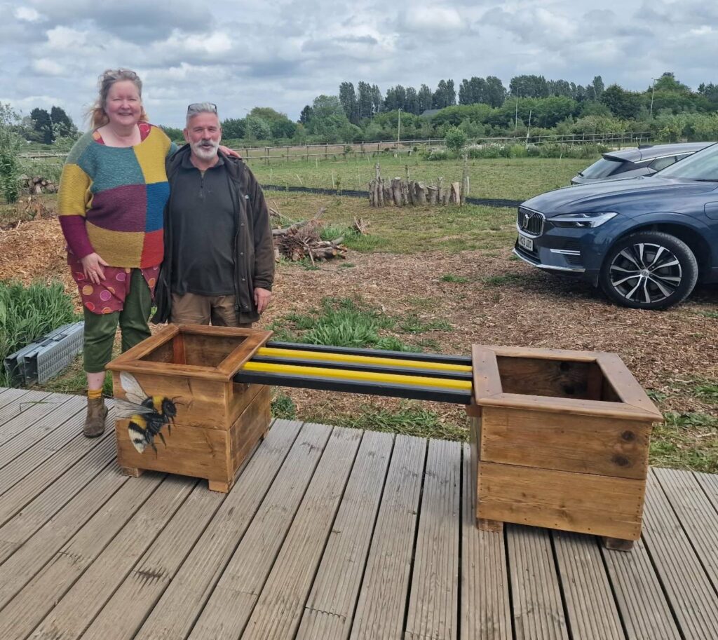 Photo of Sally Tabor presenting the handmade wooden planter and bench, with a hand-painted bee motif, to Andrew Callaghan of the Fenland Orchard Project)