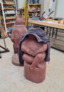 A wooden, Beatrix Potter statue of Jeremy Fisher the frog, (with a coat over it's head), waits patiently for a new base